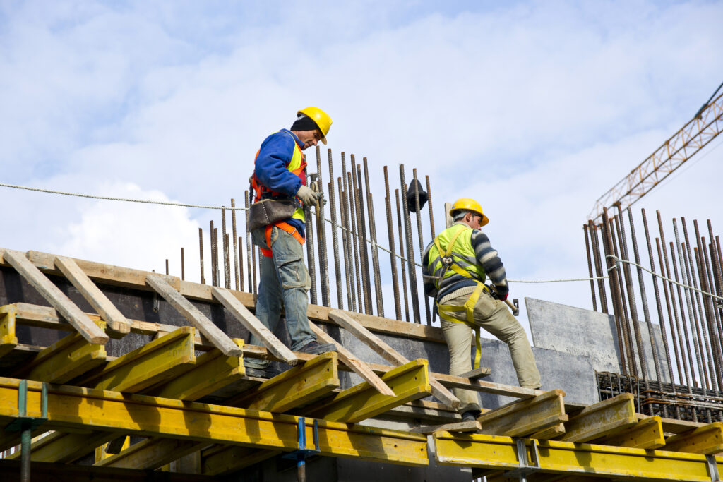 Dos trabajadores de la construcción con cascos amarillos y arneses de seguridad trabajando sobre una estructura de vigas y barras de acero.